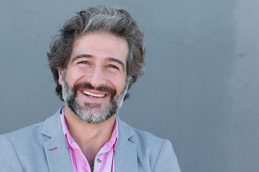 A smiling man with salt and pepper hair and beard wearing a pink shirt and gray blazer stands agains a gray wall. Get hypogonadism treatment from Morgan Burns, APRN in Central Florida.