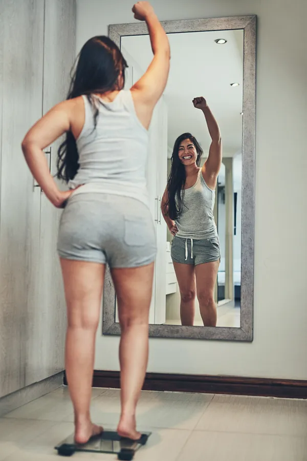 A woman in workout clothes stands on a scale looking in the mirror and pumping her hands in celebration of her successful Tirzepatide treatment from Morgan Burns, APRN in Central Florida.