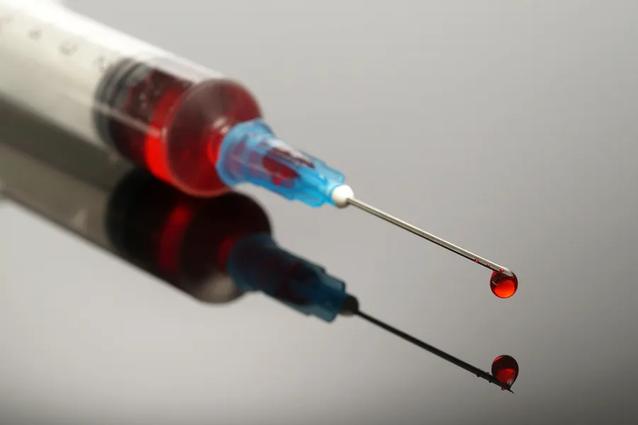 A close-up of a medical syringe filled with a red liquid, capturing a drop of the liquid falling from the needle. Lipo-B12 Weight Loss Injecitons offered by Christine Collins, MD in Westside/Beverly Hills.