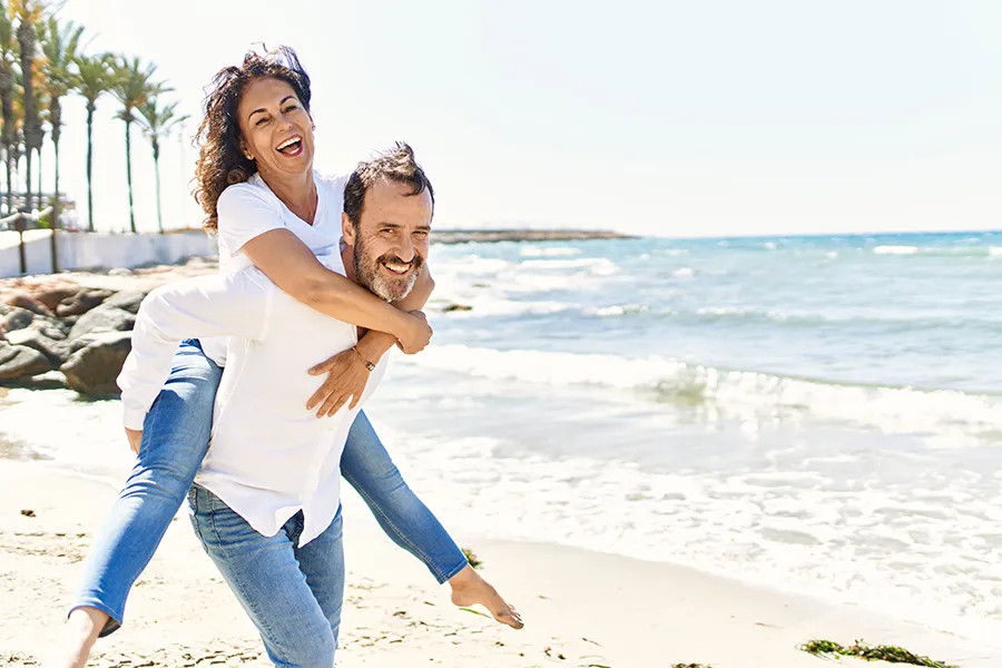 A woman piggybacks on her husband as the middle-aged couple have fun at the beach. Get NAD Therapy from Christine Collins, MD in Westside/Beverly Hills.