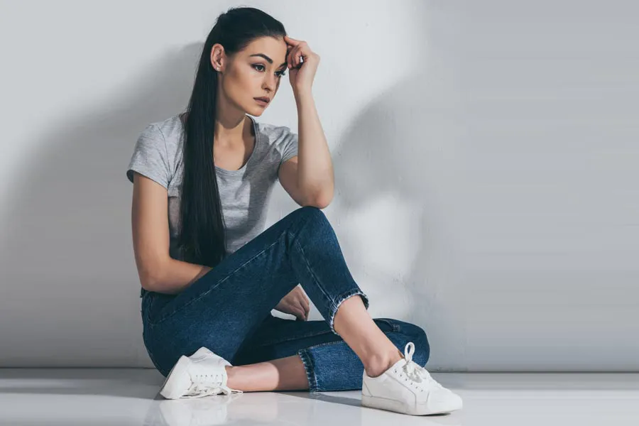 A young brunette woman in a gray t-shirt and jeans sits against a gray wall with her head resting on her hand and a distressed expression. Get help for Polycystic Ovarian Syndrome (PCOS) from Christine Collins, MD in Westside/Beverly Hills.