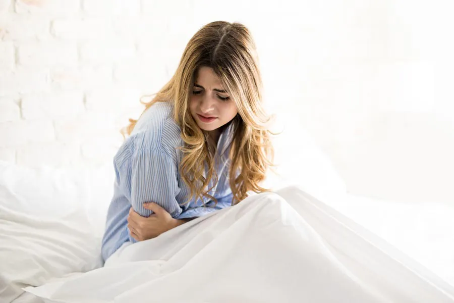 A woman with long blonde hair sits in her bed holding her stomach in pain, considering getting care for PMS from Christine Collins, MD in Westside/Beverly Hills.