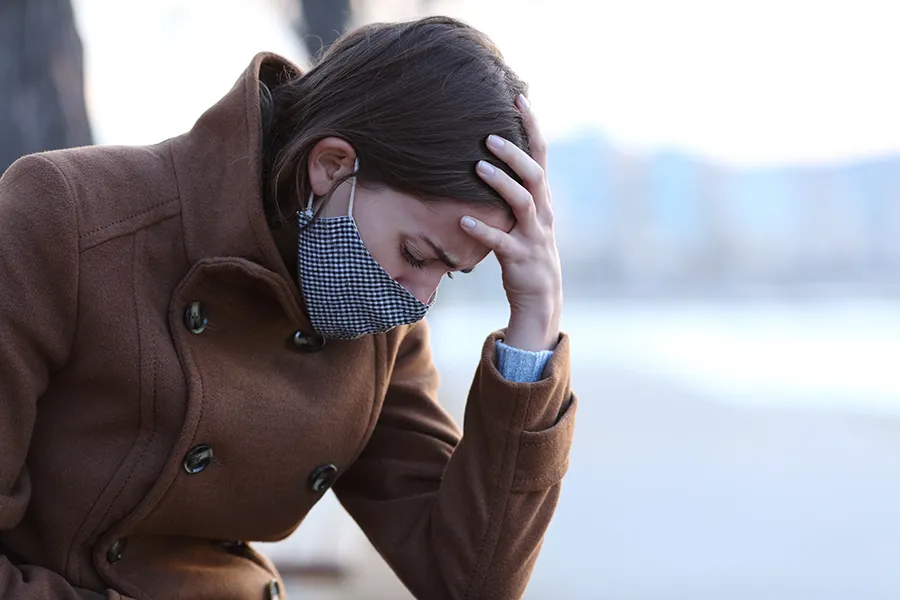 A woman in a mask sits outside with her hand on her forehead in pain. Get long covid treatment from Christine Collins, MD in Westside/Beverly Hills.