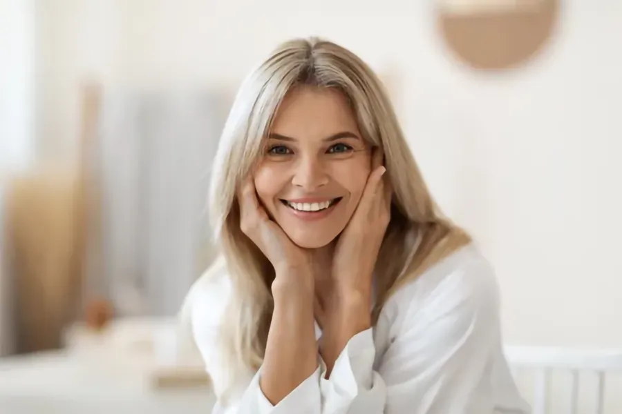 A middle-aged woman sits smiling in her living room. Schedule treatment for vaginal atrophy from Christine Collins, MD in Westside/Beverly Hills.