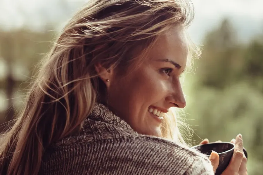 A middle-aged blonde woman in a sweater drinks tea, framed by a forest horizon. Get comprehensive women's health exams from Christine Collins, MD in Westside/Beverly Hills.