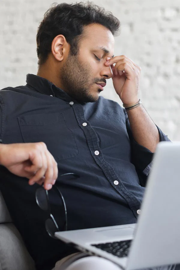 A middle-aged man with a laptop closing his eyes and rubbing his nose. He needs treatment for Adrenal Fatigue Syndrome from Frank Lanzisera, D.C. in Tampa Bay Metro.