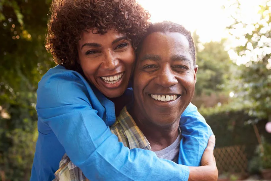 A middle-aged African American couple outside, the woman embracing the man from behind pressing her cheek to his, both are smiling due to successful Autoimmune Disorder treatment from Frank Lanzisera, D.C. in Tampa Bay Metro.