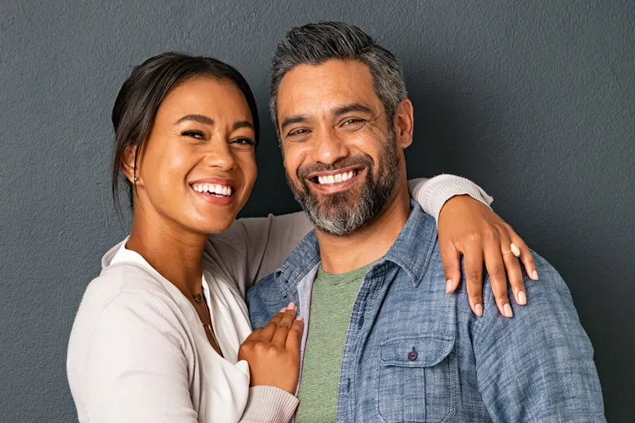 A middle-aged couple with their arms around each other standing against a gray background and smiling, happy with the bioidentical hormone therapy they received from Frank Lanzisera, D.C. in Tampa Bay Metro.