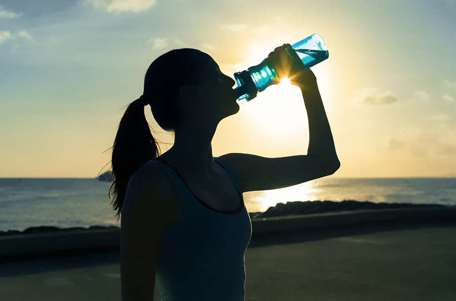 A woman in exercise clothes drinking from a water bottle on the beach, with the water and sunset in the background. Get chelation and detoxification treatment from Frank Lanzisera, D.C. in Tampa Bay Metro.