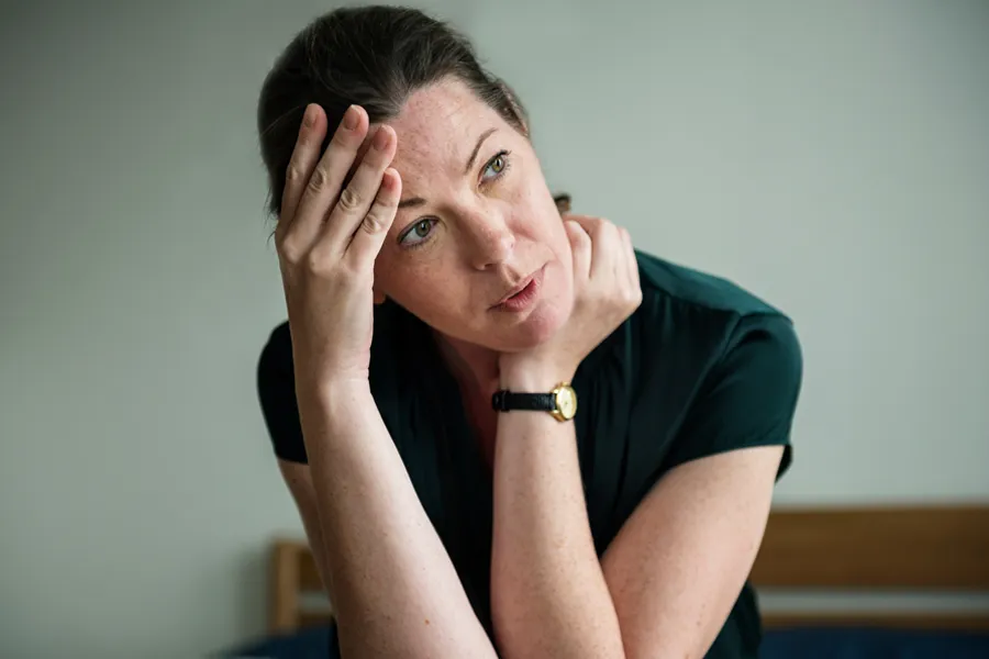 A woman in a dark blue dress sits on her bed holding her head in her hands with an exhausted expression. Get help for chronic fatigue syndrome from Frank Lanzisera, D.C. in [AERA].