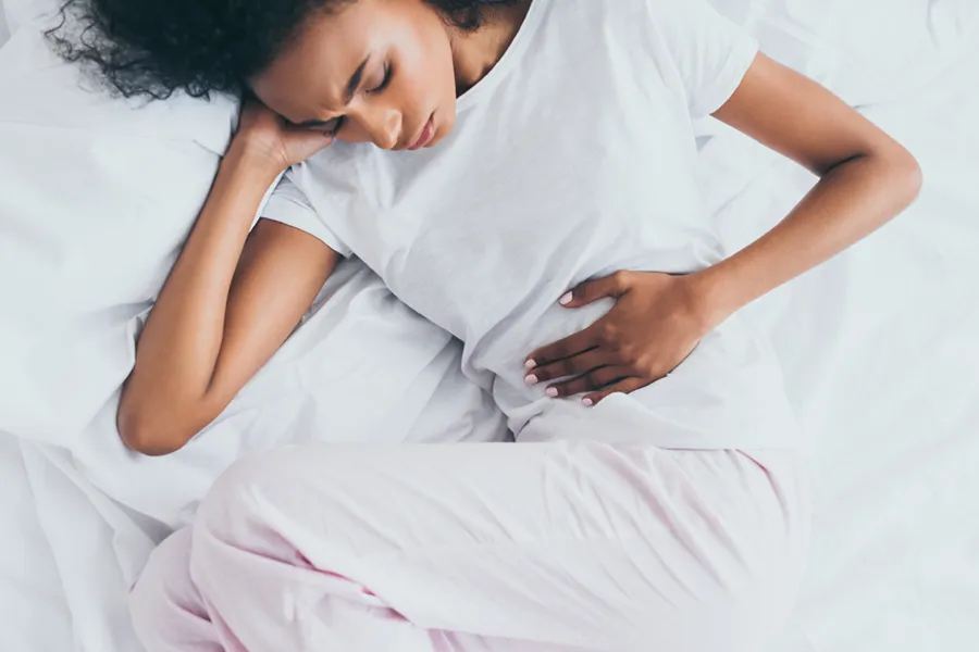 A dark-skinned woman in white clothes lays on her bed clutching her middle before getting treatment for Crohn's Disease from Frank Lanzisera, D.C. in Tampa Bay Metro.