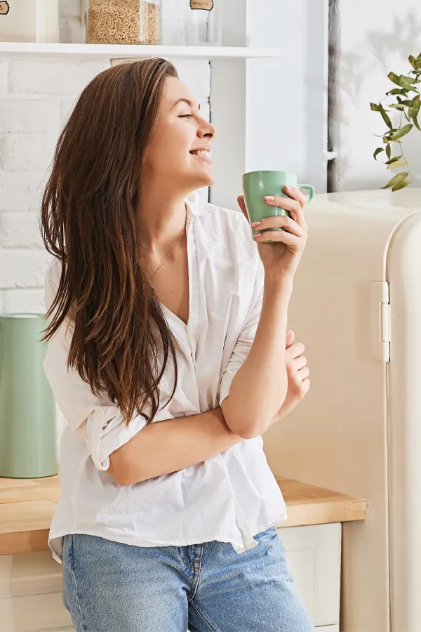 A woman with a white blouse an jeans drinking tea in her kitchen, smiling after getting treatment for Crohn's Disease from Frank Lanzisera, D.C. in Tampa Bay Metro.