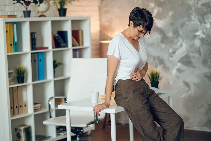 A middle-aged woman leans against her desk holing her belly in pain. Schedule IV Therapy for digestive health from Frank Lanzisera, D.C. in Tampa Bay Metro.