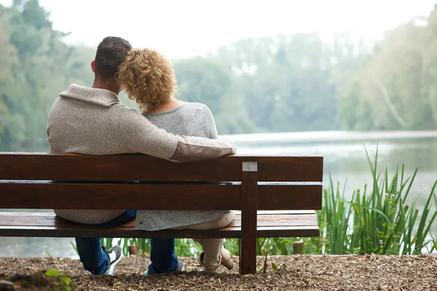 A couple wearing sweaters seen from behind sitting on a bench and looking out at a relaxing lake and forest, representing functional medicine treatment from Frank Lanzisera, D.C. in Tampa Bay Metro.