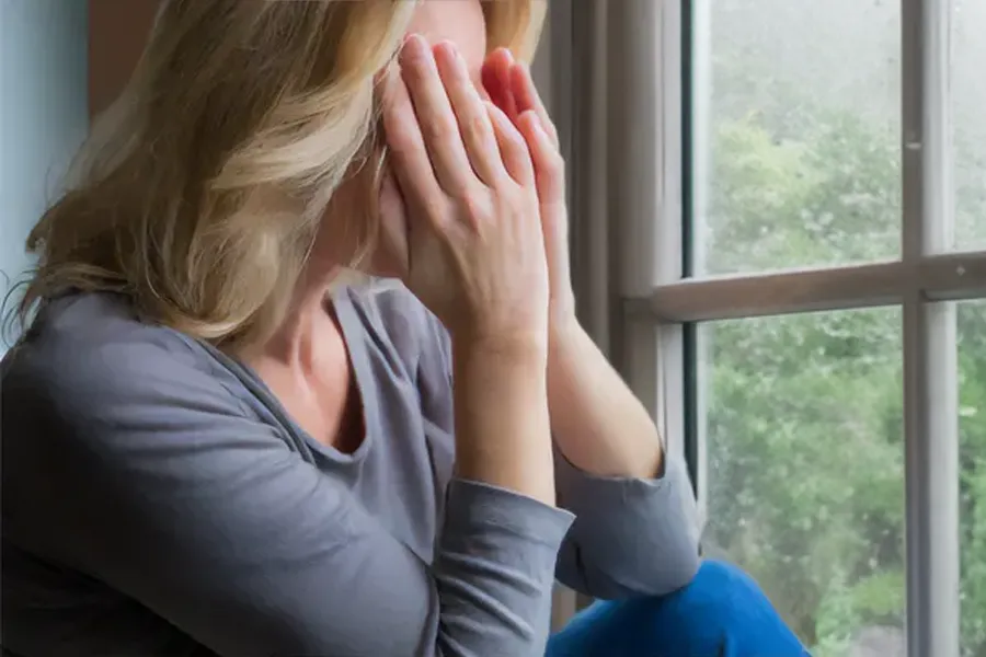 A blonde woman in a gray shirt sits in front of a window on a rainy day covering her eyes with her hands. Get treatment for Grave's Disease from Frank Lanzisera, D.C. in Tampa Bay Metro.