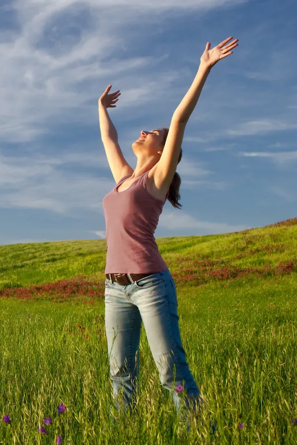 A woman in a tank top and jeans looks up with her arms outstretched to the sky in a field after successful treatment for Grave's Disease from Frank Lanzisera, D.C. in Tampa Bay Metro.