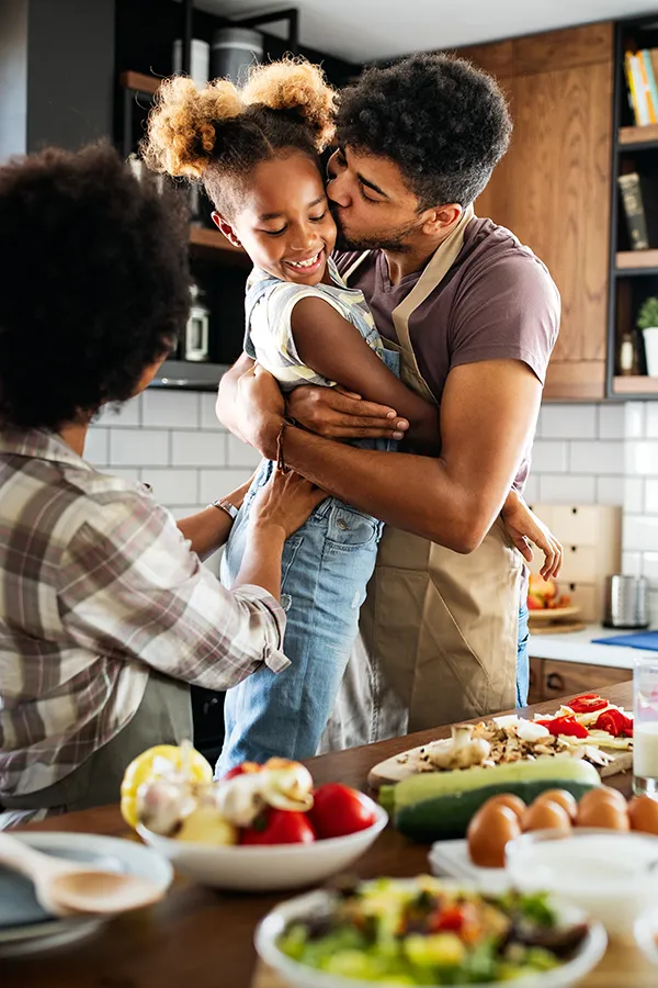 A man holds his daughter and kisses her cheek as the family makes a healthy dinner in the kitchen. Get help for digestive issues and gut health from Frank Lanzisera, D.C. in Tampa Bay Metro.