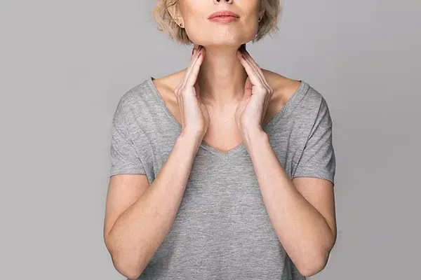 An older blonde woman in a gray t-shirt holds her hands to her neck checking for signs of thyroid problems. Get help for Hashimoto's Thyroiditis from Frank Lanzisera, D.C. in Tampa Bay Metro.