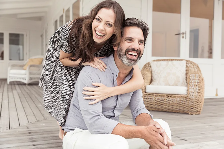A middle-aged couple on a wooden porch with wicker furniture; the woman is playfully hugging the man from behind. They are benefiting from the health and wellness services from Frank Lanzisera, D.C. in Tampa Bay Metro.