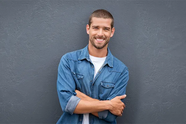 A man in a denim shirt stands smiling against a gray-blue wall, pleased with his testosterone hormone therapy from Frank Lanzisera, D.C. in Tampa Bay Metro.