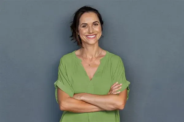A woman in a green shirt stands smiling against a gray-blue wall, pleased with her hormone therapy from Frank Lanzisera, D.C. in Tampa Bay Metro.