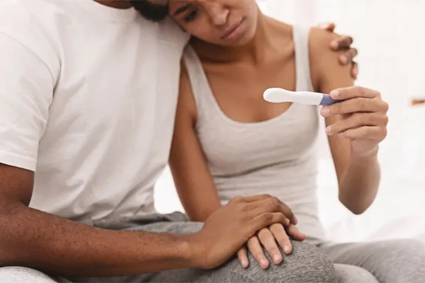 A black couple sits on a bed, disappointed with the negative results of a pregnancy test. Get treatment for infertility from Frank Lanzisera, D.C. in Tampa Bay Metro.