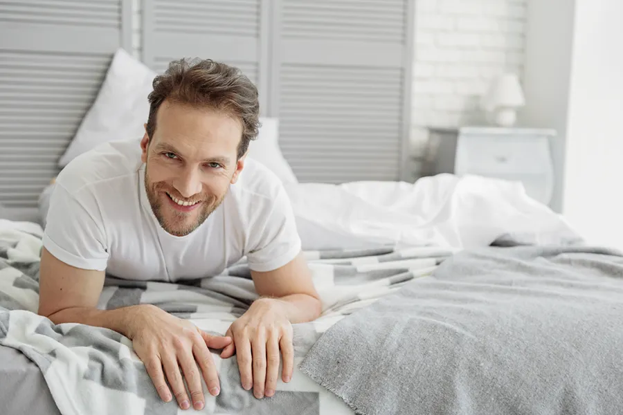 A dirty-blonde haired man lays on his stomach in bed smiling. Schedule treatment for low libido in men from Frank Lanzisera, D.C. in Tampa Bay Metro.