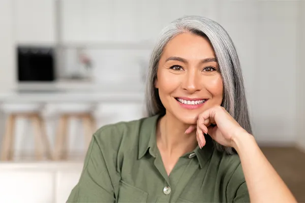 An asian woman with long gray hair and a green shirt sitting and smiling on her couch, happy with her menopause care from [NAME} in Tampa Bay Metro.
