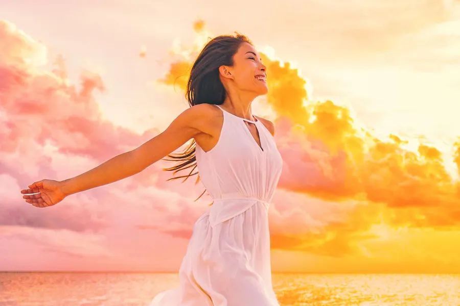 A woman in a thin white dress stretches out her arms joyously on the beach at sunrise. Get the Myers Cocktail IV treatment from Frank Lanzisera, D.C. in Tampa Bay Metro.