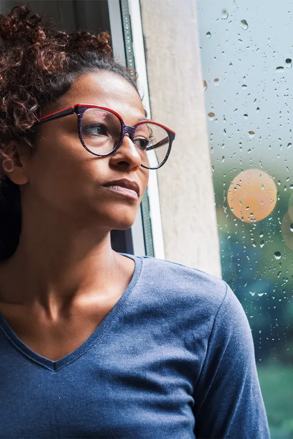A black woman with with a somber expression and glasses and a gray shirt stares out the window on a rainy day. Get treatment for Premenstrual Dysphoric Disorder from Frank Lanzisera, D.C. in Tampa Bay Metro.