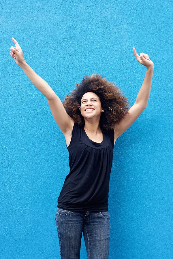 A woman in a dark blue tank top standing in front of a bright blue wall, raising her arms in celebration of relief from PMS from Frank Lanzisera, D.C. in Tampa Bay Metro.