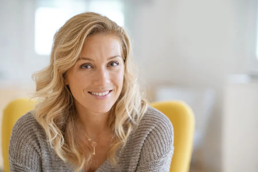 A blonde woman wearing a a gray sweater sits inside in a yellow chair, smiling and happy with her perimenopause treatment from Frank Lanzisera, D.C. in Tampa Bay Metro.