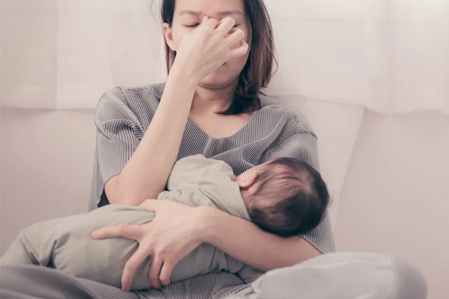 A woman nursing an infant on couch rubs her eyes with exhaustion. Get treatment for postpartum hormone imbalance from Frank Lanzisera, D.C. in Tampa Bay Metro.