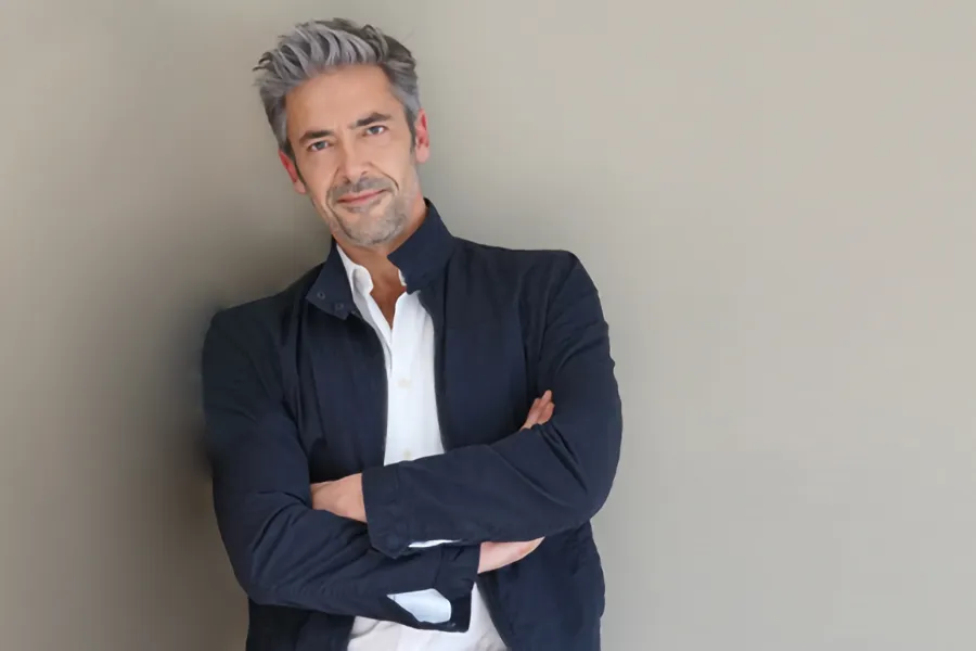 A middle-aged man in a white shirt and dark blue jacket leans against a gray wall smiling. Get testosterone for Low T from Frank Lanzisera, D.C. in Tampa Bay Metro.
